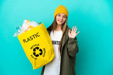 Sticker - Little girl holding a bag full of plastic bottles to recycle over isolated blue background saluting with hand with happy expression