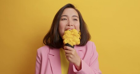 Wall Mural - Lovely Asian dark haired woman eats with appetite delicious frozen ice cream wears elegant pink jacket has happy mood isolated on yellow background. Cheerful brunette female model enjoys summer snack