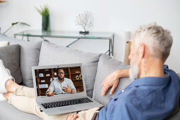 Back view over shoulder of mature man at the laptop screen with African-American man on it, two diverse male colleagues talking online, using computer app for video connection