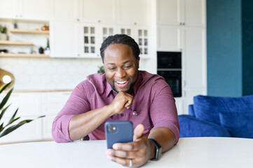 Overjoyed African-American guy enjoys spending time online in social networks, sitting at the desk at home and using smartphone, messaging and chatting, black man holds mobile and laughs
