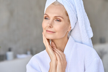 Wall Mural - Happy beautiful middle aged woman wearing bathrobe and white towel with perfect complexion touching face looking away in bathroom. Advertising of skin care spa wellness concept. Closeup portrait.