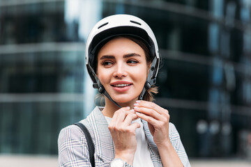 Poster - Smiling businesswoman strapping on a cycling helmet while standing in the city. Young female putting a white helmet on her head.