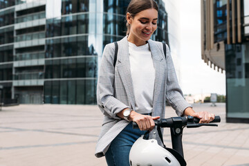 Wall Mural - Smiling blond woman holding a handlebar of an e-scooter. Female in formal wear standing on electric push scooter ready to ride.