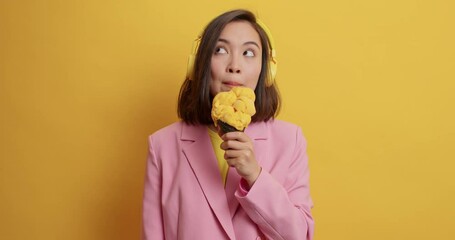 Wall Mural - Thoughtful beautiful Asian teenage girl licks ice creamduring hot summer day looks pensively at different sides listens music via headphones dressed in fashionable clothes isolated over yellow wall