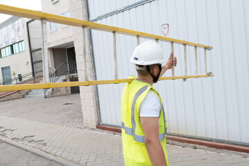 telecommunications technician walking with ladder on shoulder, wearing helmet and reflective vest.