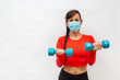 mulher fazendo exercício físico na máscara de proteção do corona vírus, com dois halteres em fundo branco.