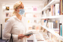 Handsome Mature Female In Protective Face Mask Choosing New Books On Shelves In Museum Shop