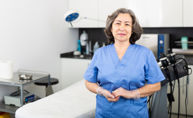 Wall Mural - Portrait of confident professional elderly woman cosmetologist wearing blue uniform standing in modern medical aesthetic office