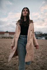 Sticker - Trendy brown haired woman wears trench and jeans on the beach with beautiful classy looking hotel on the background. Vacation on the spring on the seaside concept.