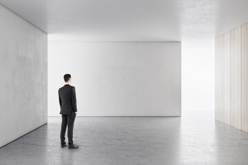 Poster - Businessman in bright concrete interior with empty wall looking at mockup place for your advertisement.