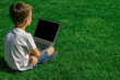 © vovan - A child sits on the grass and studies with a laptop online, enjoying nature
