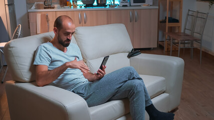 Poster - Focused adult talking with friends about social media connection using modern smartphone while sitting on couch. Tired man working late at night in kitchen using modern technology wireless