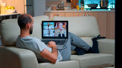 Poster - Comfortable man relaxing on couch while having online business conference with collegues using laptop computer during quarantine. Focused adult talking on videocall use webcam late at night in kitchen