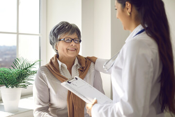 Beautiful mature woman sitting in medical office, looking at doctor and listening to advice with happy smile on face. Nurse or general practitioner reassuring senior patient during visit to hospital