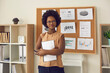 © Studio Romantic - Portrait of smart confident smiling young african american woman executive manager speech presenter with arm folded standing holding notebook near blackboard with project result report in office