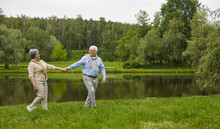 Happy Senior Couple Having A Good Time Together. Old Man And Woman Walking In A Green Park On A Fine Summer Day. Cheerful Active Grandparents Strolling Along A Grassy River Bank In The Countryside