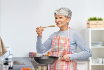 Wall Mural - cooking, culinary and old people concept - portrait of smiling senior woman in apron with frying pan and spoon tasting food over home kitchen background