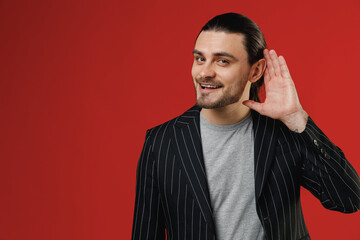 Wall Mural - Young curious nosy businessman latin man in black striped jacket grey shirt try to hear you overhear listening intently isolated on red color background studio portrait. Concept of people lifestyle