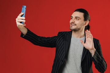 Wall Mural - Young smiling happy businessman latin man 20s with ponytail in black striped jacket grey shirt doing selfie shot on mobile phone post photo on social network waving hand isolated on red background