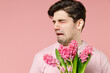 © ViDi Studio - Sick ill allergic man has red eyes runny stuffy sore nose suffer from pollen allergy symptoms hay fever hold blooming flower plant reaction on trigger isolated on pastel pink color background studio.