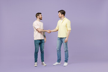 Full length two young smiling happy men friends together in casual t-shirt meeting together greet hold hands folded handshake gesture n isolated on purple background studio People lifestyle concept