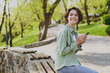 Young smiling happy student woman 20s wearing casual green jacket jeans sitting on bench in city spring park outdoors resting use mobile cell phone chat online. People active urban lifestyle concept