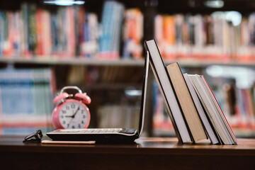 laptop computer and book, pink clock on workplace in library room with blurred bookshelf background,Education learning Concept