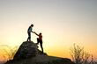 © bilanol - Man and woman hikers helping each other to climb a big stone at sunset in mountains. Couple climbing on a high rock in evening nature. Tourism, traveling and healthy lifestyle concept.