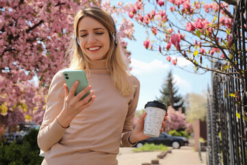 Happy woman with smartphone and coffee listening to audiobook outdoors on spring day