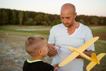 Wall Mural - Soft focus of father and son playing toy airplane in meadow at sunset with happy emotions. Family, vacation and travel concept. At sunset in summer they launch an airplane against background sky