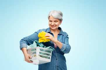 Poster - cleaning, wash and old people concept - portrait of smiling senior woman in denim shirt with towels in laundry basket over blue background