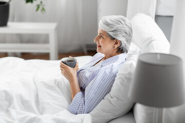 Poster - morning, old age and people concept - happy smiling senior woman in pajamas with cup of coffee sitting on bed at home bedroom