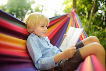 Wall Mural - Cute little blond caucasian boy reading book and having fun with multicolored hammock in backyard or outdoor playground. Summer active leisure for kids. Child relaxing in hammock.