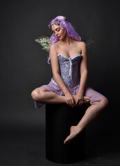 Poster - Full length portrait of a purple haired  girl wearing fantasy corset dress with fairy wings and flower crown.  Seated pose against a dark studio background.