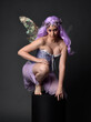 Full length portrait of a purple haired  girl wearing fantasy corset dress with fairy wings and flower crown.  Seated pose against a dark studio background.