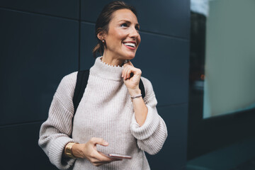 Wall Mural - Happy Caucaisan tourist using 4g wireless connection on modern cellular device, cheerful woman with digital smartphone standing at urban setting and smiling during international solo travelling