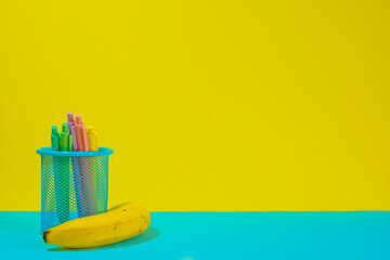 Wall Mural - Pens for back to school in a basket and a banana over a yellow and blue background. Copy space