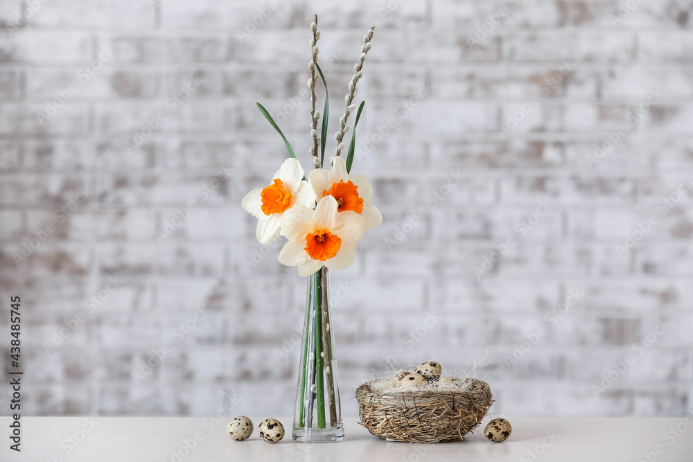 Vase with beautiful daffodils and nest with quail eggs on table near brick wall