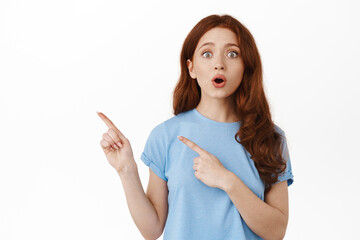 Wall Mural - Surprised and excited redhead woman telling big news, pointing fingers aside at upper left corner logo banner, gasping and say wow, impressed by advertisement, white background