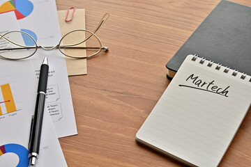 Poster - There is a note book with the word of Martech. It's on the desk with a laptop, a pen and papers of dummy graph and text.