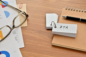 Poster - There is a word book with the word of BTA which is an abbreviation for Behavioral Targeting Advertising on the desk with papers of graphs(with dummy text), a pen and glasses.