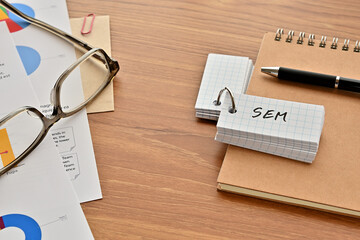 Poster - There is a word book with the word of SEM which is an abbreviation for Search Engine Marketing on the desk with papers of graphs(with dummy text), a pen and glasses.