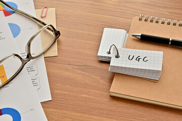 Poster - There is a word book with the word of UGC which is an abbreviation for User Generated Contents on the desk with papers of graphs(with dummy text), a pen and glasses.