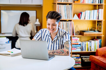 Smiling woman working on laptop while wife works behind her