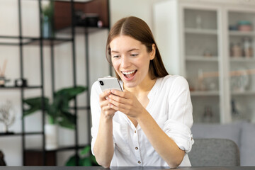 Excited happy caucasian young woman sitting at the table at home  feel euphoric checking good news on mobile phone, hiring a new job, winning online lottery