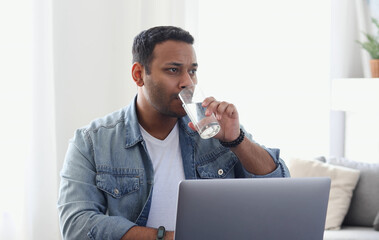 Wall Mural - Focused indian businessman working using laptop, resting and drinking refreshing water for more effective work, remote work at home