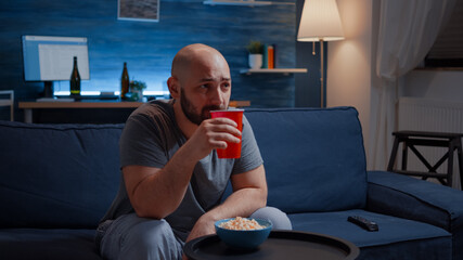 Wall Mural - Man enjoying time watching tv series at home sitting on comfortable couch dressed in pajamas eating popcorn. Excited amused home alone male drinking juice on cozy sofa in living room late night