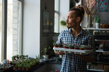 Wall Mural - Handsome young man with plants standing by the window at home