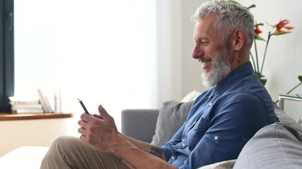 Cheerful and healthy grey-haired middle aged hipster man in casual shirt using smartphone sitting on the sofa at home, mature guy enjoys scrolling news feed, looks at the screen and smiles, side view
