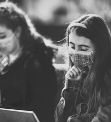 Sticker - Two young girls seated on a bench in the city park using tablets wearing masks in pandemic time